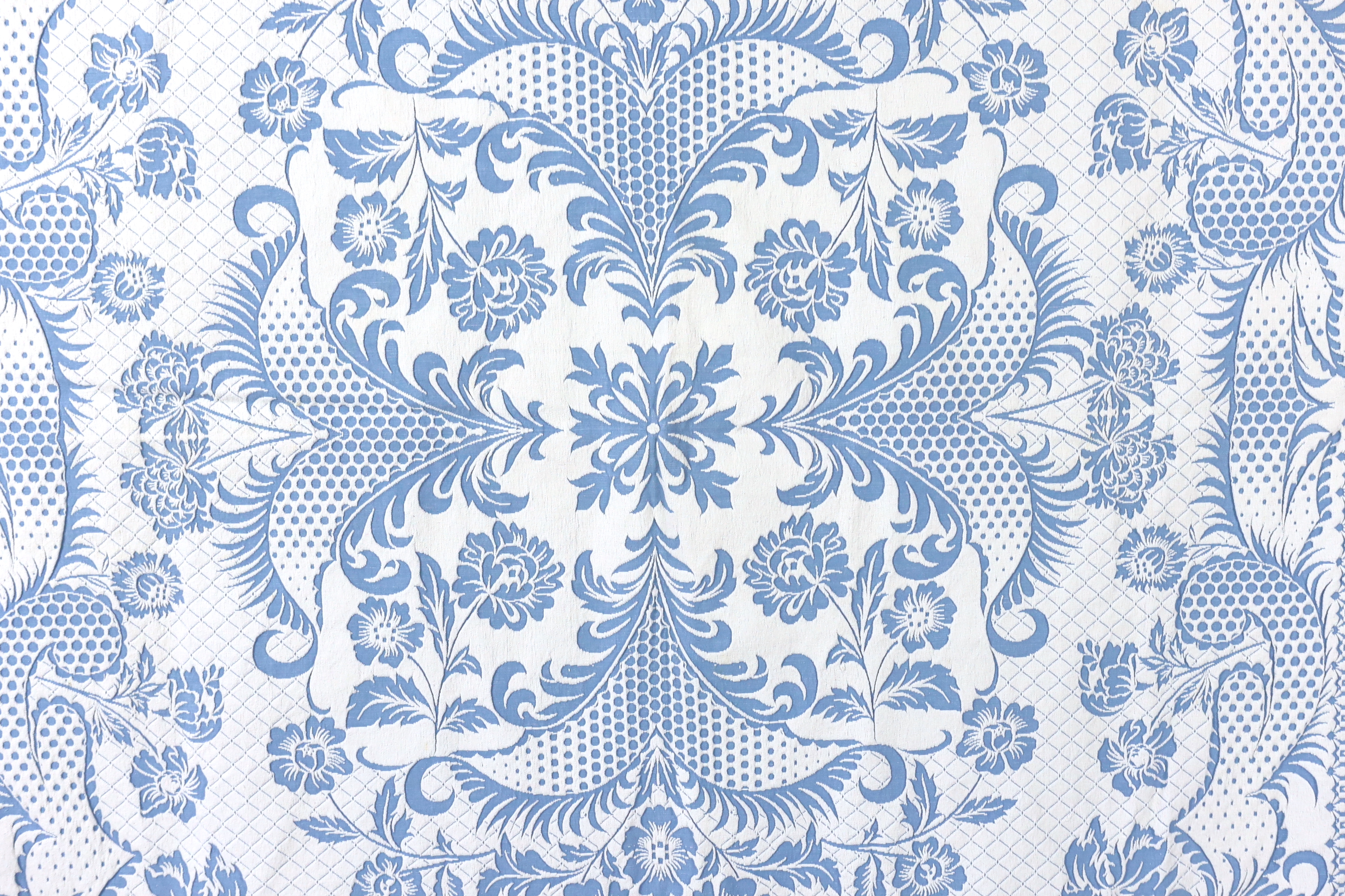 An early 20th century woven 'Marcella' style bed cover, with a blue ground and a white raised floral central cartouche and a wide patterned border, bed cover 194cm wide x 225cm long